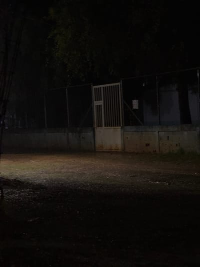 The image depicts a dimly lit, possibly rainy, outdoor scene in El Bajío, Venezuela. A section of a fence, likely chain-link, runs vertically across the image. Adjacent to the fence is a concrete structure with one or more access panels or doors. The ground appears to be wet asphalt or pavement. Dense, dark foliage is visible on the left side of the fence. Power lines or cables are visible in the lower left corner. No people are visible.