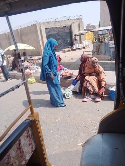 The image depicts an outdoor market scene in Chiranchi, Nigeria, viewed from inside a vehicle, with parts of its yellow frame and a light-colored seat visible in the foreground.
In the midground, a female subject stands left-center, clad in a full-length light blue hijab and dark sandals, holding a small red object. To her right, two female subjects are seated on a low concrete barrier; one wears a dark brown hijab, and the other, to her right, wears a light orange/beige hijab and pink sandals. A metal platter containing pinkish items is on the ground between the standing and seated women. A blue plastic bucket and folded fabric are positioned near the rightmost seated woman.
In the background-left, several male figures are engaged in market activities under a yellow umbrella, with various goods displayed on tables and the ground. A tall, light-colored concrete wall topped with coiled barbed wire extends across the background, featuring a large, dark metal gate. Beyond the wall on the right, a multi-story, light-colored building and several makeshift market stalls with corrugated metal roofs are visible. The ground in the market area appears dusty and unpaved, while the immediate foreground consists of a paved road surface. The sky is clear and bright.