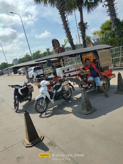 A man in a red vest and blue shorts sits on the side of a red, open-air vehicle that is likely a tuk-tuk or similar local transport. He is leaning against the vehicle and smiling at the camera. The vehicle has a canopy and a metal frame. In front of the vehicle are two motorcycles, one white and one black. There are also several traffic cones on the ground. In the background, there are palm trees, a white car, and a fence. The scene appears to be in a dusty, outdoor area with a paved surface. The watermark indicates the photo was taken in Bavet Quarter, Cambodia, on June 24, 2026, at 09:18.