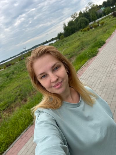 A young woman with blonde hair and freckles is taking a selfie outdoors. She is wearing a light blue sweatshirt. The background shows a grassy area, a paved path, a body of water, and trees. The sky is cloudy. The image appears to be taken in Breytovo, Russia, as indicated by the presence of a lamppost with a Cyrillic sign and the general landscape which is consistent with the region.