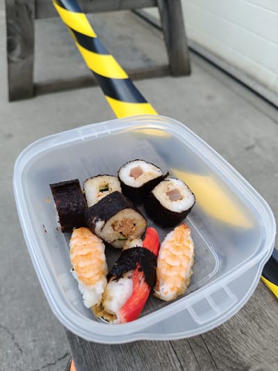 The image shows a clear plastic container filled with various types of sushi. Among the sushi are pieces with shrimp, tuna, and what appears to be imitation crab. Some rolls are maki-style, wrapped in seaweed, while others are nigiri, with a topping over rice. The container is placed on a wooden surface, possibly a table or bench, outdoors.

In the background, there are wooden posts and a yellow and black striped barrier, suggesting an area that might be under construction or has some safety precautions in place. The ground appears to be concrete. The lighting suggests it is daytime, possibly overcast as there are no strong shadows. The scene is casual, with the sushi likely prepared for a simple meal or snack. There is no visible text in the image. The setting appears to be Târgu Jiu, Romania.