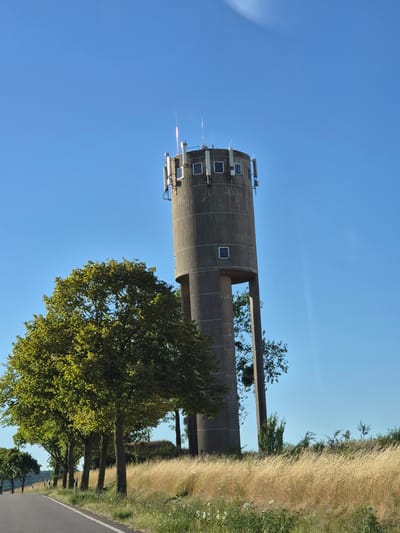 The image shows a concrete water tower situated in an open, rural landscape. The tower is cylindrical with a textured, gray exterior. It is topped with several antennas and what appear to be communication equipment, along with a few small, square windows. There are also two large, rectangular openings in the upper portion of the tower, revealing its interior structure and allowing trees to be seen through them.
Surrounding the water tower are several mature trees with lush green foliage. A paved road runs along the right side of the image, bordered by a grassy embankment covered in dry, golden grass. The sky is a clear, vibrant blue, indicating a sunny day. The lighting suggests it is likely late afternoon or early evening, as evidenced by the warm tones in the dry grass and the long shadows that might be present but are not clearly visible in this rotated image. The scene conveys a sense of tranquility and isolation, characteristic of a rural setting. There are no people visible in the image. The location is identified as Schengen, Luxembourg.