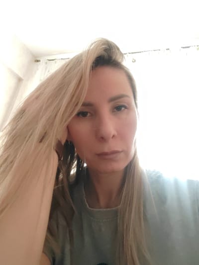 A woman with blonde hair is looking directly at the camera with a neutral expression. Her hair is partially up and falls to her shoulders on either side of her face. She is wearing a grey t-shirt. Her left hand is touching her face. The background appears to be a brightly lit room, possibly near a window with light-colored curtains. The location is Италмас, Russia.