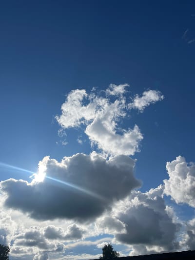 The image shows a bright blue sky filled with dramatic, fluffy clouds. The sun is peeking through a large, dark grey cloud on the left side of the frame, casting a brilliant lens flare across the sky. The clouds are varied in size and texture, with some appearing bright white and others shadowed. The lower portion of the image shows a dark silhouette of trees and a rooftop, suggesting an outdoor scene in an urban or suburban environment. The scene appears to be set in Серебрянский, Russia, as indicated.