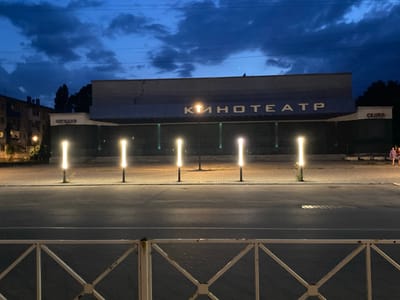 The image depicts a street scene at dusk in Antratsyt, Ukraine, featuring a building with Cyrillic lettering that reads "КИНОТЕАТР" (KinoTeatr), meaning cinema. The building's facade is a light gray with a blue band across the top, and it is illuminated by several tall, vertical lights positioned along the sidewalk in front of it. The street in the foreground is asphalt, with white lane markings. A white metal fence with a diamond pattern is visible in the bottom portion of the image. In the background, a multi-story apartment building is visible to the left, and trees line the right side of the frame. The sky is a deep blue with dark, dramatic clouds. Two individuals are walking away from the camera on the sidewalk near the right edge of the frame.