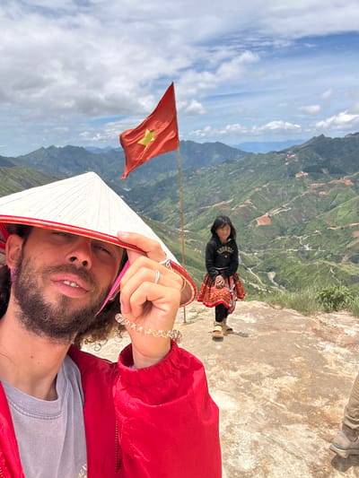 A selfie taken outdoors shows a person wearing a conical hat and a red jacket, holding a clear spiral bracelet and a ring on their left hand. Behind them, a young girl in a black hoodie and a colorful skirt stands on a rocky surface. In the background, a Vietnamese flag with a yellow star on a red field waves in the wind, set against a dramatic landscape of rolling green hills and mountains with terraced fields. The sky is partly cloudy. The location is identified as Bắc Yên, Vietnam.