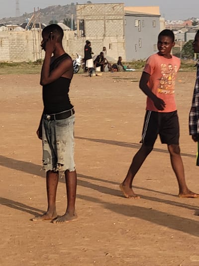 Two barefoot young males are observed on a dirt-covered ground surface. One male is wearing a dark-colored tank top, tattered jean shorts, and a belt, while the other male is wearing a light-colored t-shirt and dark-colored shorts. In the background, buildings and other people are present. This is the Ibo area of Nigeria.