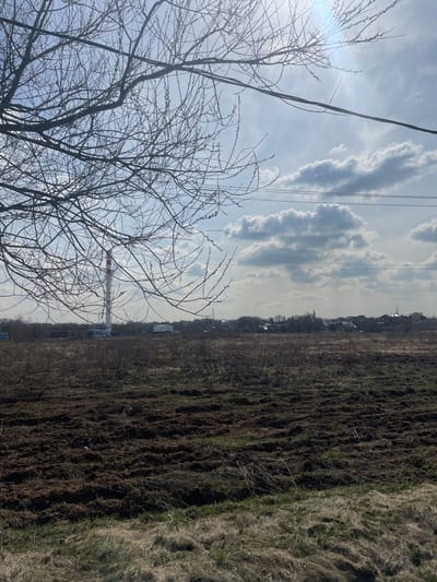 The image shows a field in Fabriki 1 maya, Russia, next to a bare tree in early spring. The sky is partly cloudy, with a mix of blue sky and white clouds. Power lines run across the sky. In the background, there are buildings, including a tall chimney with red and white stripes. The field is brown and appears to have been recently tilled. Patches of dead grass can be seen.