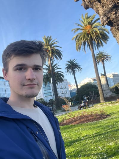 The image captures a daylight scene in a public park within the city of La Coruña, Spain. In the immediate foreground, a young adult male is depicted from the chest up, taking a selfie. He has short brown hair, light facial stubble on his chin and jawline, and wears a dark blue zipped jacket over a light cream or beige long-sleeved shirt. He looks directly at the camera with a neutral expression.
The background features a vibrant green lawn liberally dotted with small white flowers, suggesting daisies. Multiple tall palm trees, characterized by long trunks, green fronds, and clusters of orange dates, rise against a clear, bright blue sky. Beyond the park's greenery, several modern multi-story buildings with numerous windows are visible. To the right, a substantial portion of a dark, gnarled tree trunk with rough bark is present at the edge of the frame. Further into the park, in the mid-ground, a stone monument or pillar is partially visible, and several indistinct individuals are observed seated on park benches or walking. The scene is illuminated by strong natural sunlight, casting discernible shadows.