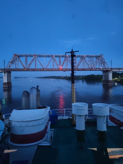 The image depicts a view from a boat, likely on a river, at dusk or dawn. In the background, there is a large, steel truss bridge with railway tracks. The river reflects the light and the bridge, showing a red light on the bridge. The shoreline is visible in the distance with green trees. In the foreground, part of the boat is visible, including white and red structures, potentially covers or vents.
It is not possible to confirm the location in the city of Tarkanovo, Russia from the given image.