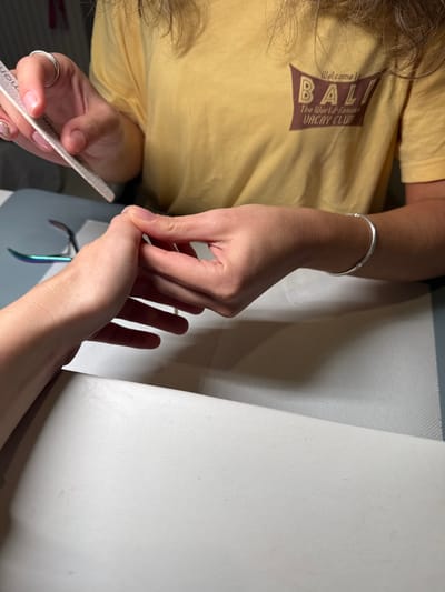 A person in a yellow "Bali Vacay Club" t-shirt files another person's nails. The person filing the nails wears a silver ring and bracelet. The person receiving the manicure has their hand spread out on a white surface. Manicure tools are also visible, including a nail file and nail clippers.