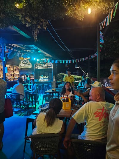 The image depicts a lively outdoor scene at a bar or restaurant, likely in Ban Klong Haeng, Thailand, at night. The setting is illuminated by vibrant blue and green lights, creating a festive atmosphere. Strings of small international flags are draped across the overhead structure, suggesting a diverse clientele or a celebration of global unity.

Several people are visible, engaged in various activities. In the foreground, a man in a light-colored t-shirt with a graphic print is looking down at a tablet. Behind him, a woman in a white top is seated, and another individual is partially visible. Further back, several other patrons are scattered throughout the seating area, some appearing to be dining or socializing. The bar area is lined with shelves stocked with bottles, and two prominent Jägermeister signs are displayed, indicating the establishment's offerings.

The overall mood seems relaxed and social, with people enjoying their evening. The time of day is clearly night, evidenced by the artificial lighting and the dark sky. The presence of lush greenery on the left side of the image suggests an outdoor, perhaps tropical, environment. Visible text includes "Jägermeister" on the signs and "NEW YORK LOS ANGELES LONDON" on the man's t-shirt.