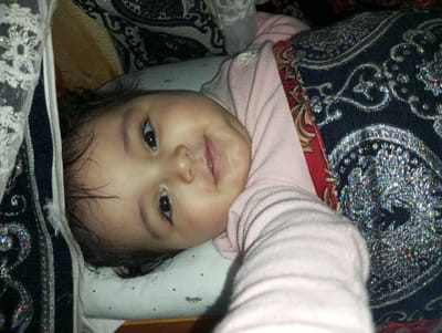 A photographic image captured in Galaosiyo, Uzbekistan, depicts a young human subject, approximately infant to toddler age, positioned supine with the head slightly tilted. The subject possesses dark hair, dark irises, and fair skin, exhibiting an upward curve of the lips consistent with a smile. The subject is dressed in a light pink, ribbed, long-sleeved garment. The subject lies on a white fabric surface adorned with small dark dots and faint outlined figures. To the immediate left of the subject's head (viewer's left), a white lace material with reflective sequins is present. To the subject's right (viewer's right), a dark textile embellished with reflective silver paisley-like patterns is draped, partially overlapping a red fabric featuring gold and black designs. A portion of the subject's right arm extends towards the foreground of the image. The immediate environment comprises textiles of varying patterns and textures.