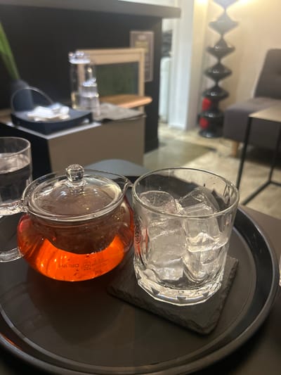 The image presents a close-up, eye-level view of a beverage service on a dark, circular serving tray within an indoor establishment in 마포구, South Korea. In the foreground, a transparent glass teapot, labeled "HARIO," is filled with an amber-orange liquid, likely tea, with an internal infuser visible. Adjacent to the teapot on the right is a clear, textured glass tumbler containing numerous large ice cubes, resting on a dark, rectangular coaster. A third clear glass, filled with water, is partially visible on the far left of the tray.
The background is intentionally out of focus, depicting a softly lit interior space. To the left, a dark surface displays indistinct items including a water pitcher, a small dark box with a white object, and a light-framed item. To the right, a tall, black floor lamp with a segmented, hourglass-like design emits a warm light, illuminating a section of a grey upholstered chair and a slender-framed side table. The floor appears to be a light, possibly concrete, material. No individuals are present in the frame.