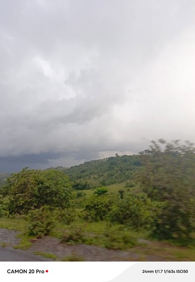 A cloudy sky hangs over a lush, rolling green landscape, likely the city of Nguroje, Nigeria. The foreground is dominated by a blurred image of the terrain, suggesting movement, possibly from a vehicle. Dark, heavy clouds gather on the left side of the sky, hinting at impending weather. The middle ground features a hillside covered in dense green vegetation and scattered trees. The far background is a hazy expanse of sky and distant hills. Text at the bottom of the image reads "CAMON 20 Pro" and "24mm f/1.7 1/163s ISO50." No people or specific objects are clearly identifiable due to the motion blur.