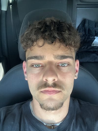 A selfie-style photograph depicts a young man with light skin, light brown curly hair, and stubble. He has light-colored eyes. He wears a dark t-shirt and a silver chain necklace. He appears to be seated in a car, as evidenced by the car seat and interior trim visible. A jacket is visible in the upper left. The photo is taken at an angle, likely from above. The location of the image, Ravnishte, Bulgaria, is only known because of the prompt instructions and is not visually supported by the image.