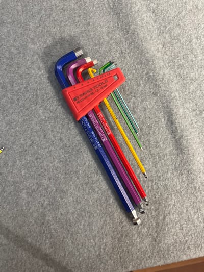 A set of PB Swiss Tools Allen wrenches is arranged on a gray textured surface. The Allen wrenches are color-coded and arranged from largest to smallest, with sizes indicated by numbers etched into the plastic holder. The largest wrench, colored blue, appears to be 10 mm, followed by purple (8 mm), red (6 mm), yellow (5 mm), green (4 mm), light green (3 mm), light blue (2.5 mm), and finally a smaller size not clearly visible. The red plastic holder has the brand name "PB SWISS TOOLS" and product information "PB 212.LH-10 RB : 1,5 - 10 mm" printed on it. The Allen wrenches are L-shaped, with one end of each tool bent at a 90-degree angle. The background is a plain gray surface with a slight texture, suggesting a fabric or carpeted material. There are no people or other objects present in the image. The image's origin is Cologny, Switzerland.