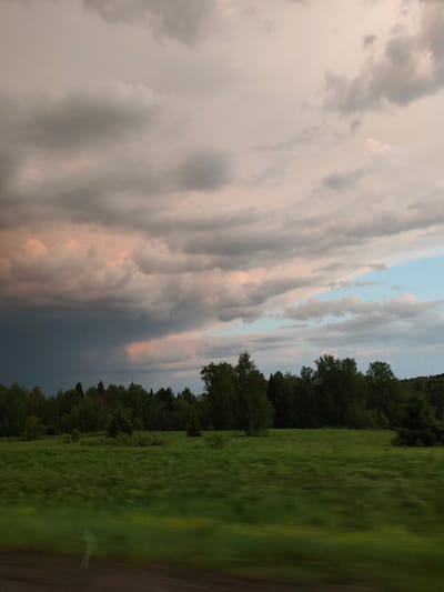 The image shows a rural scene under a dramatic sky. Dark storm clouds are gathering on the left, while the right side of the sky has lighter clouds tinged with pink and blue. Below the clouds, a line of trees forms the horizon, varying in height and density. A green field stretches out in the foreground, showing some blur likely due to movement. The lower edge of the image is dark, possibly indicating the edge of a road or the inside of a vehicle.
Without additional information, it is not possible to confirm that this picture was taken in Glazunovo, Russia.