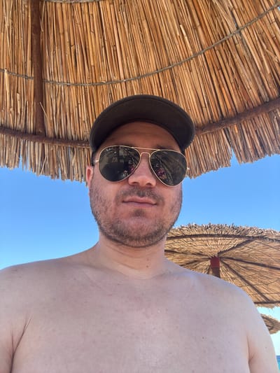 A man wearing a dark baseball cap and aviator sunglasses takes a selfie from a low angle. He is shirtless and his shoulders and upper chest are visible. The background is a bright blue sky with two thatched beach umbrellas. The umbrella directly above the man is large and woven from dry reeds. The second umbrella, in the background, is smaller and appears to be identical in style. The scene suggests a sunny day at a beach in Toroni Municipal Unit, Greece, likely a tourist area.