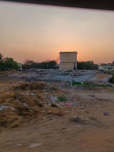 The image displays an outdoor scene in Kothacheruvu, India, at either dawn or dusk, characterized by an orange and pale blue gradient sky. The immediate foreground is blurred, composed of reddish-brown soil, a mound of dried fibrous material, and scattered multi-colored plastic litter. A weathered, yellow-painted cylindrical water storage tank with a metal access ladder on its left side is centrally located, positioned atop a rocky elevation. A white pipe extends downwards from the tank, partially obscured by rocks. Stacks of light-colored bricks are visible near the base of the tank. The midground and background feature dry vegetation, including bushy green trees and shrubs. To the far left, a building with yellow and blue sections is partially visible through trees. To the far right, a light-colored building with a green roof is discernible, accompanied by utility poles and overhead wires. No individuals or discernible actions are present. A dark horizontal band is visible across the top of the frame.