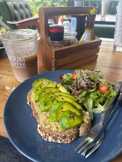 A slightly tilted, close-up shot depicts a meal and drink on a wooden table. On a dark blue plate, there is avocado toast with a side salad. The avocado is sliced and sprinkled with black seeds. The salad contains lettuce, cucumber, and tomato. Silverware rests on the plate next to the salad. To the left of the plate is a clear plastic cup with a lid and straw, containing a beverage, labeled "Premium Coffee Koh Phangan". Behind the drink is a wooden condiment caddy containing various jars and bottles. Through a window in the background, greenery and light are visible. The scene is likely set in Baan Kai, Thailand, based on the "Premium Coffee Koh Phangan" label on the cup, and a casual dining environment.