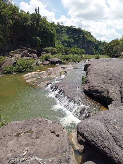 The image captures a view of a shallow river flowing over rocks, creating small cascades. The river is surrounded by lush green vegetation and large, dark rocks. In the background, a steep, rocky cliff covered in dense trees rises majestically. The sky is partly cloudy with a few white clouds against a blue backdrop. A person wearing a yellow shirt and a red hat is visible on the right side of the image, standing near the edge of the rocks. The scene appears to be a natural landscape, possibly a waterfall or river within a park or wilderness area. The specific location in Tân Phú, Vietnam, suggests it is a natural attraction within that region.