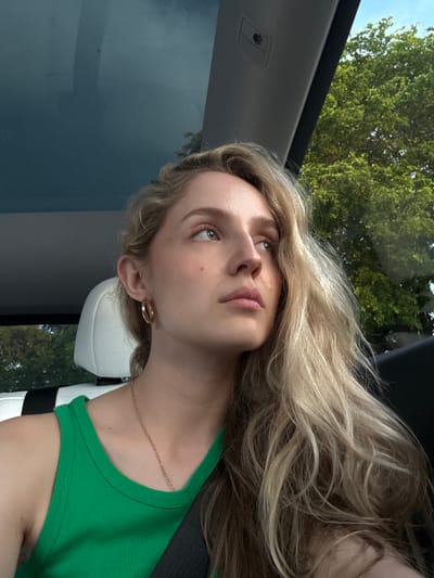 A young woman with blonde, wavy hair is seen from the chest up, looking to her right. She is wearing a green tank top and a gold necklace. She has large hoop earrings and her seatbelt is visible across her chest. The background shows the interior of a car with a sunroof and a view of green trees and a blue sky outside.