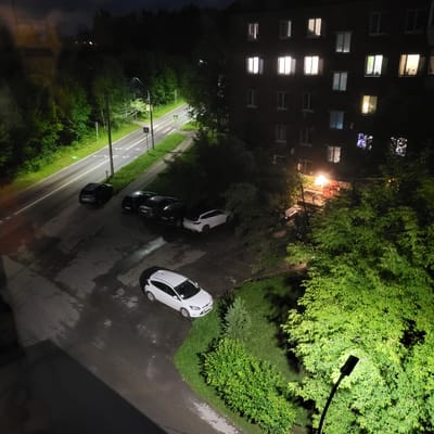 The image depicts a night scene in a residential area, likely a courtyard, with an apartment building on the right and a road on the left. The apartment building is dark, with several windows illuminated from within. Trees and foliage are present throughout the scene, some of which are illuminated by a nearby streetlight. A road with visible lane markings runs alongside the building, and there are several cars parked in a lot adjacent to the road. One white car is prominently parked in the foreground, while several dark-colored cars are parked in a row further back. The ground appears wet in places, suggesting recent rain. The overall impression is that of a quiet night in a Russian residential district, possibly in the village of Глебовский, Russia, given the context. There are no people visible in the image.