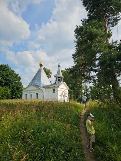 In the village of Ulitkino, Russia, a white Orthodox church with a golden dome and a bell tower stands on a grassy hill. A dirt path leads away from the church, bordered by tall grass and trees. A young boy in a yellow shirt and a blue cap walks down the path, facing away from the camera. Further up the path, a man in a green t-shirt and shorts walks towards the church. The sky is blue with scattered white clouds.