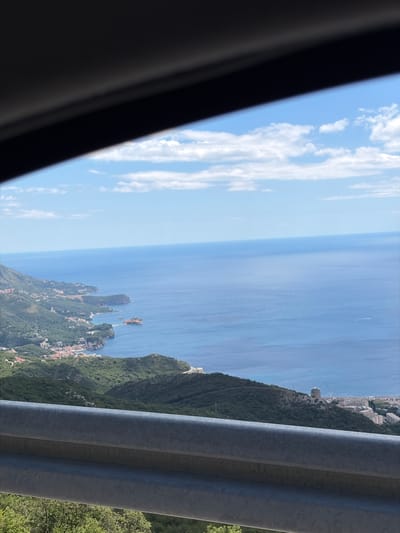 An aerial, eye-level, medium shot captures a scenic coastal view of Stanišići, Montenegro, on a sunny day. The foreground is dominated by a dark, shadowy object partially obscuring the top of the frame, possibly the interior of a vehicle. Below this, a silver metal guardrail runs horizontally across the lower portion of the image. Beyond the guardrail, a lush green hillside slopes down towards the coast. A small village with red-roofed buildings is nestled along the shoreline. The blue Adriatic Sea stretches out to the horizon, meeting a light blue sky dotted with white, fluffy clouds. The coastline is characterized by rocky cliffs and peninsulas jutting into the water. In the distance, on a hill, a white building with a distinctive tower is visible. The overall impression is a tranquil and picturesque landscape.