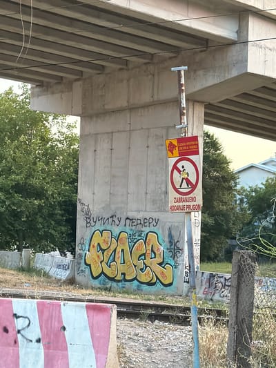 The image captures a concrete pillar supporting an elevated structure, likely a bridge or overpass, in Burtaiši, Montenegro. The pillar is covered in graffiti, with a large yellow and orange tag prominently displayed. To the right of the graffiti, a sign with red and yellow elements is attached to a rusty metal pole. This sign features a red circle with a diagonal line through it, containing a pictogram of a person walking, indicating a prohibition. Below this, text in Cyrillic reads "ZABRANJENO HODANJE PRUGOM," translating to "Walking on the tracks is forbidden." Another smaller sign above the prohibition sign displays the logo of "Željeznička Infrastruktura Crne Gore AD - Podgorica," the Montenegrin railway infrastructure company. The surrounding environment shows railway tracks, overgrown vegetation, and some concrete barriers. The overall scene suggests an area near a railway line that is not intended for pedestrian access.