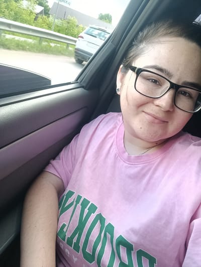 A young Caucasian woman with dark hair, fair skin, and freckles is taking a selfie in a car. She is wearing black-rimmed glasses and a pink t-shirt with green lettering that reads "BROOKLYN." She has a silver chain necklace on. She is smiling and looking at the camera. The background shows the interior of the car, the window, and a blurry view of the outside with a road, a guardrail, trees, and a white car. The image appears to have been taken on a cloudy day.