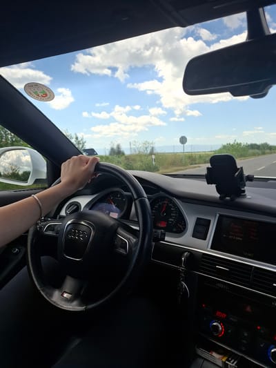 A person is driving an Audi, likely an S-Line model given the steering wheel badge, on a road near Brezovo, Bulgaria. The environment is sunny with a blue sky and scattered clouds. The car's interior features include a dashboard displaying a temperature of 20.0 degrees Celsius, radio tuning to 87.5 MHz (Bulgaria), a navigation system (or infotainment screen), and a cell phone holder mounted on the dashboard. A sticker, likely related to a local municipality, is visible on the windshield near the rearview mirror. The driver's hand is on the steering wheel, adorned with a bracelet. The speedometer indicates a speed that is hard to distinguish, and the tachometer shows low RPM.