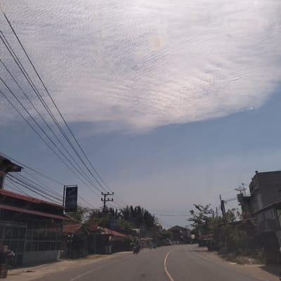 The image depicts a street scene in Cibrektunong, Indonesia, on a day with thin, high clouds filling the sky. A two-lane road with a yellow center line curves slightly to the right. The road is flanked by buildings and vegetation. The buildings appear to be a mix of commercial and residential structures, some with red tile roofs. Power lines crisscross overhead. A sign is visible on the left side of the road, bearing the letters "LA" above smaller text. A stop sign is also visible. A few motorcycles can be seen on the road. The overall impression is of a typical street view in a small Indonesian town on a sunny day.