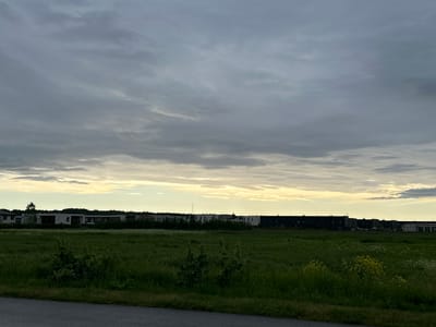 The image depicts an outdoor scene in Harkujärve, Estonia, characterized by a broad expanse of green grassland in the foreground, leading towards a line of modern buildings in the mid-ground. The grassland is dense and appears somewhat overgrown, with scattered patches of wildflowers, some yellow and white, visible amongst the green. The buildings, a mix of residential and possibly commercial structures, are low-rise and contemporary in design, featuring clean lines and a variety of facades, including white, grey, and dark-colored panels. A dark grey asphalt path borders the bottom of the image, suggesting a road or walkway. The sky above is overcast with heavy, grey clouds, but a band of lighter, yellowish light is visible along the horizon, indicating either dawn or dusk. The overall impression is one of a developing or established suburban or peri-urban area under a diffused, muted sky. There are no people or animals visible in the frame.