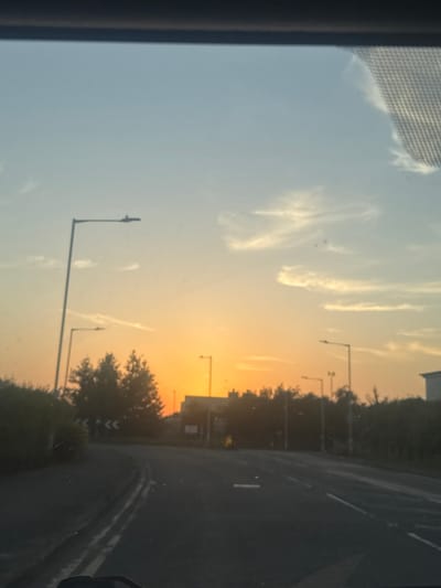 The image is a vertical photograph taken from inside a vehicle, looking out through the windshield. The scene depicts a road with streetlights lining the right side, bordered by trees. The sky is a prominent feature, transitioning from a clear pale blue on the left to a warm orange and yellow gradient on the right, indicative of a sunset. Wispy clouds are scattered across the sky, catching the light of the setting sun.
The activity taking place is travel, suggested by the perspective from inside a car and the presence of a road. The time of day is clearly sunset. No people are visible, but there are dark silhouettes of trees and bushes. The streetlights suggest an urban or suburban setting, likely a road in the Borough of Swale, United Kingdom, as per the provided context. The overall mood is serene, characterized by the soft colors of the sunset and the quiet road. There is no visible text in the image.