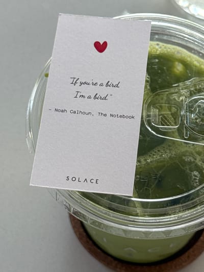 A clear plastic cup filled with a green beverage, possibly matcha, is displayed. A white card with a red heart sits on top of the cup. It features the quote "If you're a bird, I'm a bird." attributed to Noah Calhoun from "The Notebook." The word "SOLACE" is printed in simple, sans-serif font at the bottom right corner of the card. The cup rests on a brown coaster.