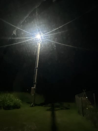 This is a nighttime, low-light photograph taken outdoors, likely in a rural or semi-rural setting within the city of Мясоцкі сельскі Савет, Belarus. The primary subject is a tall, utility pole with a bright light fixture at its apex, casting an intense glare that creates prominent starburst-like light streaks radiating outwards into the dark sky. The pole appears to be made of metal and has a rectangular electrical box mounted on its lower section. A long, dark shadow of the pole is cast onto the green grass below. To the left of the pole, there is an area of dense, green vegetation. To the right and further in the background, a chain-link fence with a wooden post is visible, partially obscured by darkness. The ground is covered in green grass, and the overall atmosphere is dark, illuminated solely by the artificial light source. There are no people or animals visible in the image.