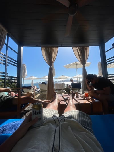 The image captures a scene from a shaded cabana in Cabo San Lucas, Mexico, overlooking the ocean. Two individuals are visible: one sitting and leaning over a table with drinks, and another closer to the viewer, holding a can of Modelo beer. Outside the cabana, there are several beach umbrellas and lounge chairs. The environment is sunny with clear blue skies, typical of Cabo San Lucas. The foreground features the interior of the cabana, including wooden structures and a ceiling fan.