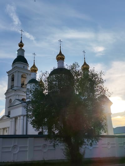 The image shows a white Orthodox church with golden domes, characteristic of Russian architecture. The church is partially obscured by a tree in the foreground. The sky is blue with some clouds, and the sun appears to be setting, casting a warm light. The church has a bell tower and multiple onion domes topped with crosses. A white decorative wall runs in front of the church. The location is Faliukovo, Russia.
