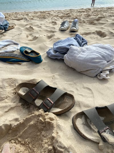 The image shows a sandy beach, with a blue towel lying partially on the sand and two pairs of sandals. The sandals are brown and made of leather. One pair is lying on the sand, the other is partially buried in the sand. The towel is folded and slightly rumpled. The image was taken near the coordinates (latitude: 28.694, longitude: -14.017), which is in the beach area near El Cotillo in La Oliva, Fuerteventura, Spain.  The scene suggests the beach is a popular spot for relaxation.  The image suggests a person has left their belongings on the beach while enjoying a swim or another activity.