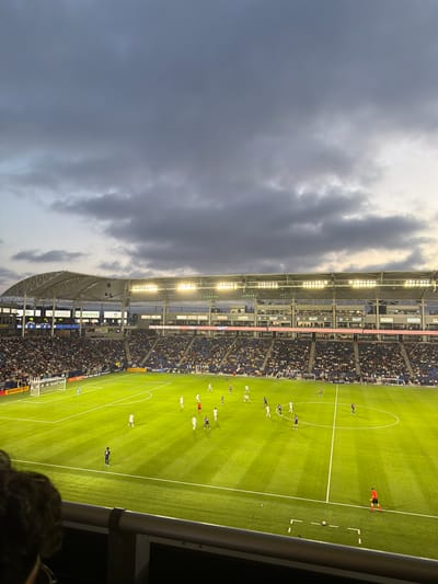 The image shows a soccer game being played at Dignity Health Sports Park in Carson, California, United States. The sky is overcast with dark clouds. Stadium lights illuminate the field. Spectators fill the stands. Soccer players are in action on the field.