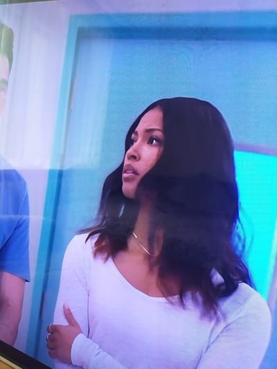 The image shows a medium shot of two people in an indoor setting, likely a room with a bright blue wall in the background. On the right, a woman with long, dark hair is looking to her left. She is wearing a white long-sleeved shirt and a delicate necklace. Her left arm is bent, and her left hand rests on her right arm, displaying a few rings. On the left of the image, partially visible, is a person wearing a blue t-shirt. The lighting appears to be bright and even. There is no indication of the city of Turka, Ukraine, in the image.