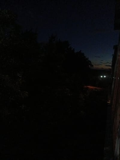 The image depicts a dark, possibly twilight or night scene with a dimly lit outdoor environment. In the foreground and left side of the frame, dense foliage of trees is visible, appearing as dark silhouettes against a darker sky. The sky itself is a deep navy blue, with a hint of lighter colors near the horizon on the right, suggesting the last vestiges of daylight or distant city lights.
Further into the scene, on the right side of the image, the edge of a building is visible. It has a rough, textured surface, possibly brick or concrete, with vertical lines that suggest construction or weathering. Adjacent to this building, a road or path can be inferred, with a warm, orange-red glow emanating from it, possibly from streetlights or a vehicle. Two bright, white headlights of a vehicle are also visible in the distance, moving away from the viewer. A few other faint, distant lights are also present.
The overall impression is one of a quiet, possibly suburban or semi-rural area on the outskirts of a city, illuminated by the remaining ambient light of the sky and artificial light sources. The location is specified as Kuznetsk, Russia. There are no people clearly visible in the frame.