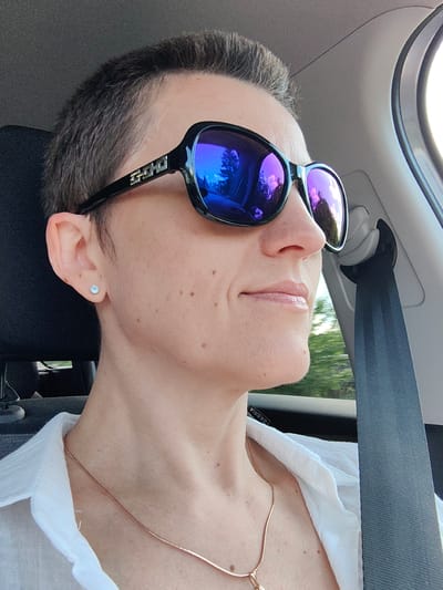 A woman with short, dark hair and sunglasses is pictured from the chest up, looking to the right. She is wearing a white collared shirt and a rose gold necklace. A blue seatbelt is visible to her right. The background shows trees and a bright sky, suggesting the scene takes place outdoors during the daytime, possibly while traveling in a vehicle. The exact location of Ilyanskiy selskiy Sovet, Belarus cannot be determined from the visual information provided.