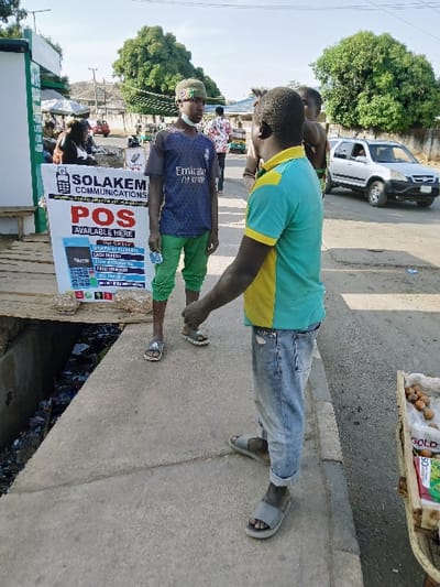 Two individuals stand on a sidewalk next to a drainage ditch filled with refuse. One person wears a blue and green shirt with a yellow collar and light-colored jeans. The second person has on a blue shirt with "Emirates" printed on it and green pants. They are standing next to a sign advertising "SOLAKEM Communications POS available here," depicting a handheld device with various services listed. In the background, there are other people, cars, and a fruit stand. A lush green tree is visible in the distance.