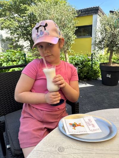 A young girl wearing a pink t-shirt and a pink baseball cap with a Minnie Mouse design sits at an outdoor table. She is holding a milkshake in a clear glass with a straw. On the table, to her right, is a silver tray with a white napkin folded on it. The napkin has a cartoon image of an ice cream cone with a face, and text printed on it. The girl is looking at the camera with a slight smile. The setting appears to be a cafe or restaurant with tables and chairs, and greenery, including an olive tree, in the background. A yellow building is visible in the distance. The sunlight suggests it is daytime. The napkin has the text "LUST AUF EIS!" printed on it, which translates to "Fancy some ice cream!" from German. This suggests the location is likely in Germany.