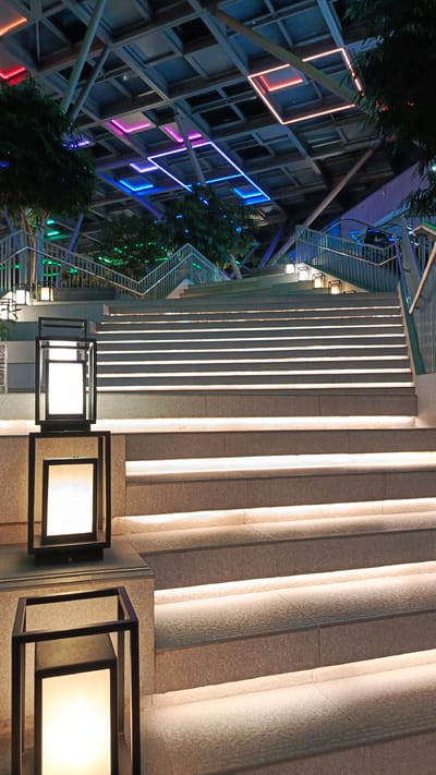 The image depicts an outdoor modern staircase illuminated by embedded lights on each step, leading upwards towards a ceiling structure. The ceiling features a grid of square panels, some of which are illuminated with colorful neon lights in shades of blue, pink, and orange. Trees with dark green foliage are visible on either side of the staircase and under the ceiling structure. In the foreground, two modern, black-framed lanterns with glowing white light are placed on a stone ledge to the left of the stairs. The overall scene suggests a contemporary architectural design, likely in a public space like an atrium or walkway, at dusk or night, given the artificial lighting. The provided context of Wanasari, Indonesia, is noted but not visually evident from the image itself.