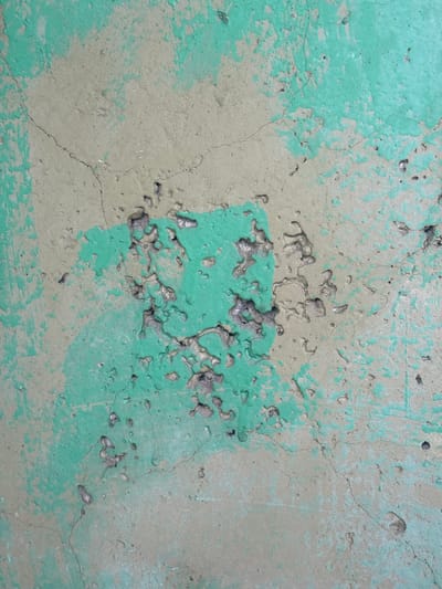 The image shows a close-up of a weathered, textured concrete surface with patches of faded green paint. The concrete exhibits numerous small pits, cracks, and areas where the paint has chipped away, revealing the underlying grey material. The surface appears to be outdoors, as indicated by the natural wear and tear. There are no people or specific objects visible that would place this image within a particular city or location, including Vyksa, Russia. The image focuses solely on the abstract texture and condition of the concrete.