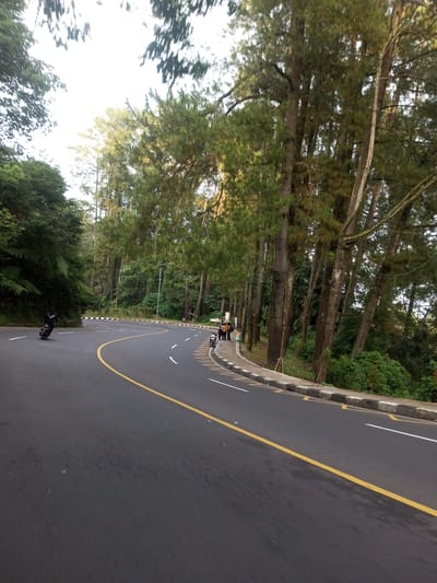The image depicts a curved road in Taunggyi, Myanmar. The road is asphalt, marked with a solid yellow line and broken white lines. A person on a motorbike is seen riding around the curve. On the roadside, several people are standing near parked motorcycles. Trees line the road, with dense foliage visible. The roadside has a curb with alternating white and black markings. A street lamp is visible.